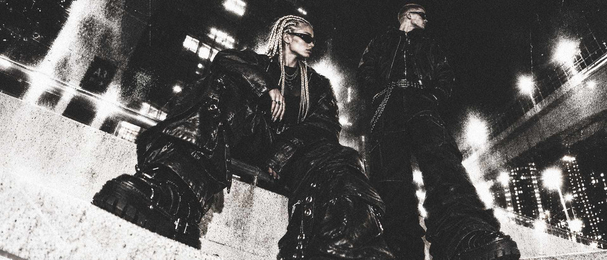 Two models in black jackets, wide-leg pants, chunky boots and sunglasses on concrete steps at night, photographed in black and white.