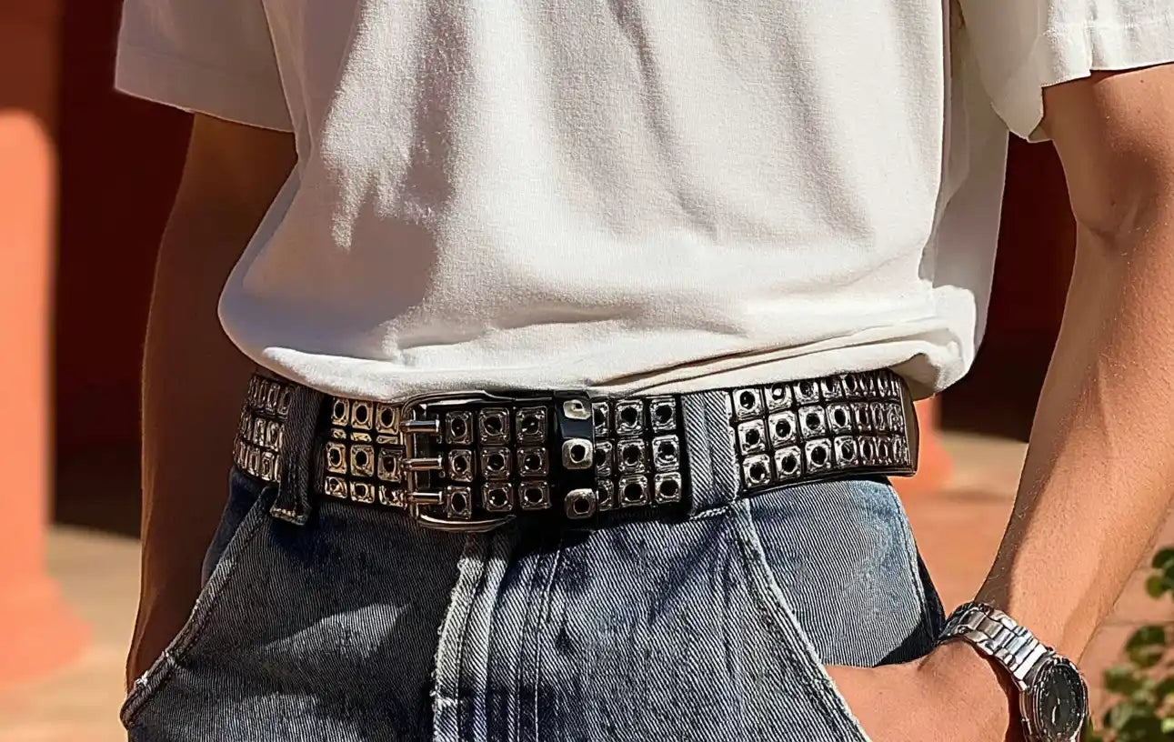 Black studded streetwear belt with rows of silver grommets worn over washed blue jeans.